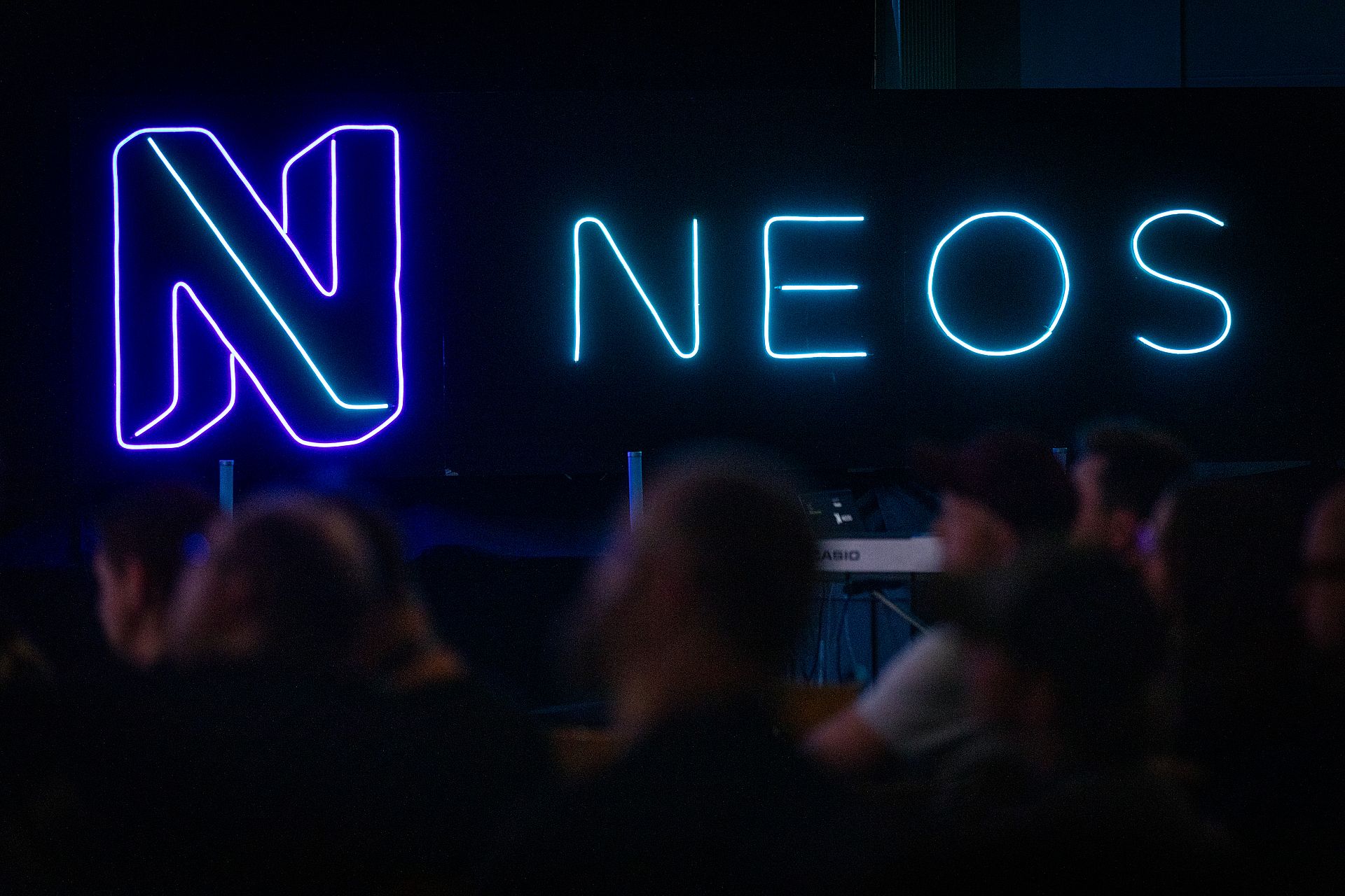 Der Neos-Schriftzug leuchtet in einem dunklen Raum mit Personen